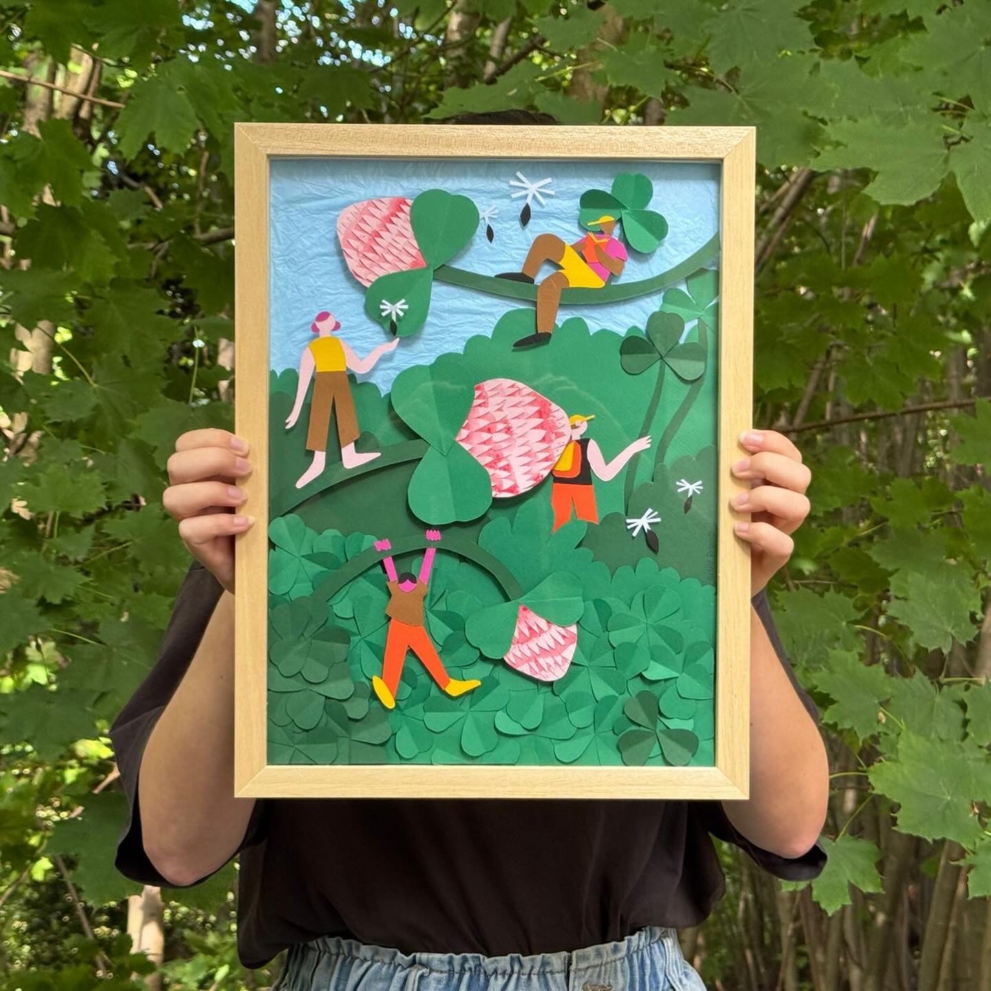Une personne qui tient un cadre dans lequel il y a un oeuvre en papier découpé qui représente des personnages et des trèfles.