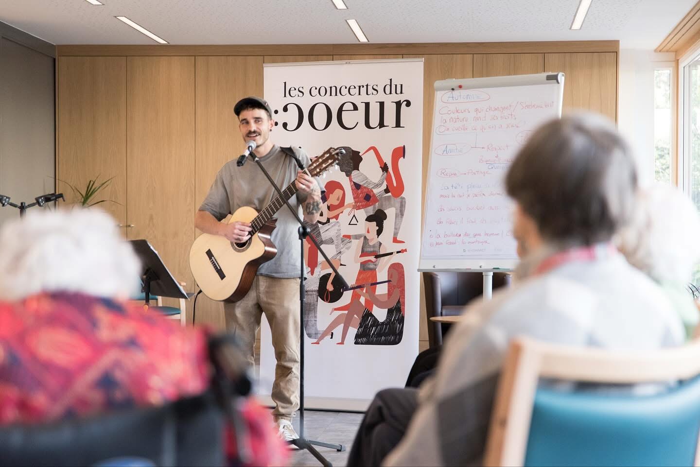 Photo d'un concert donné dans un home pour personnes âgée où l'on voit 2 personnes âgée au premier plan et au centre de l'image un homme qui hante et qui joue de la guitare. Derrière lui un rollup avec l'illustration créée pour les concerts du Coeur en Valais