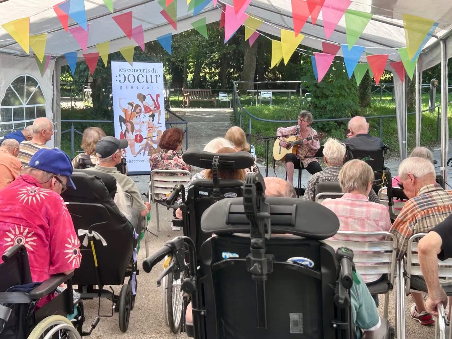 Photo d'un concert donné dans les jardin d'un home pour personnes âgées. Au premier pan des personnes en fauteuils roulants. Une personne assise chante et joue de la guitare. Au fond on voit de rollup avec l'illustration des concerts du coeur de genève
